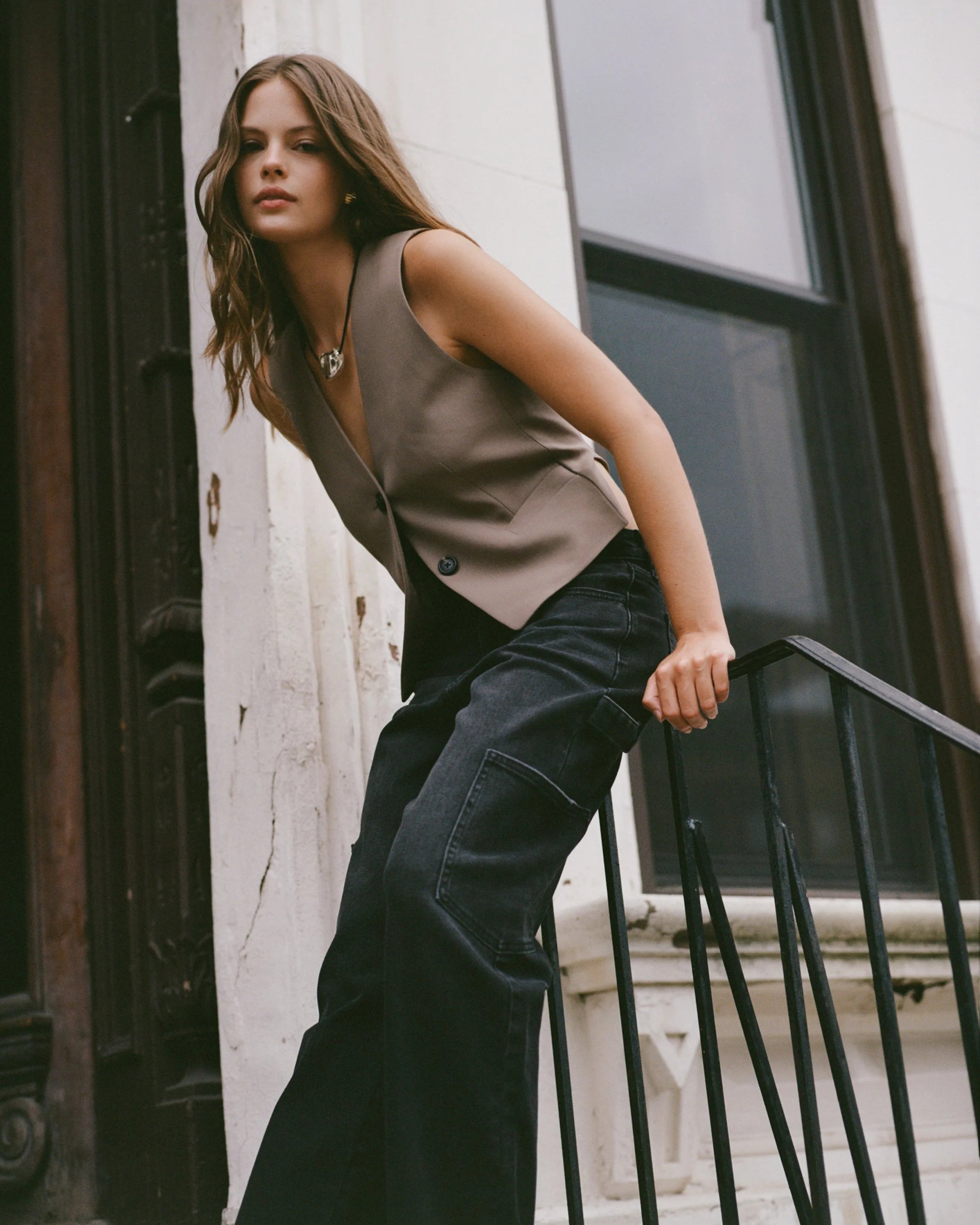 Woman leaning against a building wall wearing a sleeveless top and dark pants.