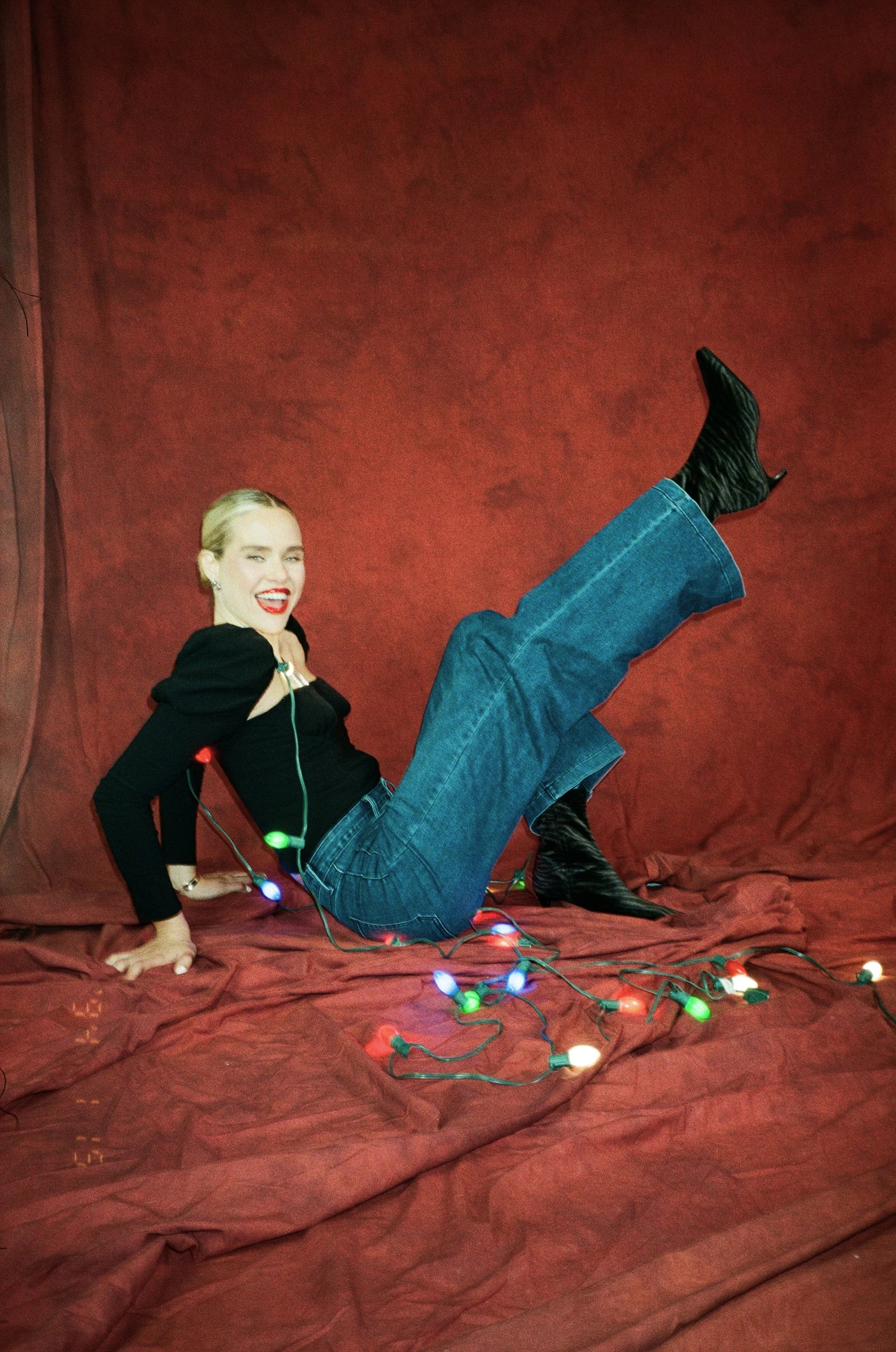 Person lying on a red fabric with Christmas lights, against a red curtain background