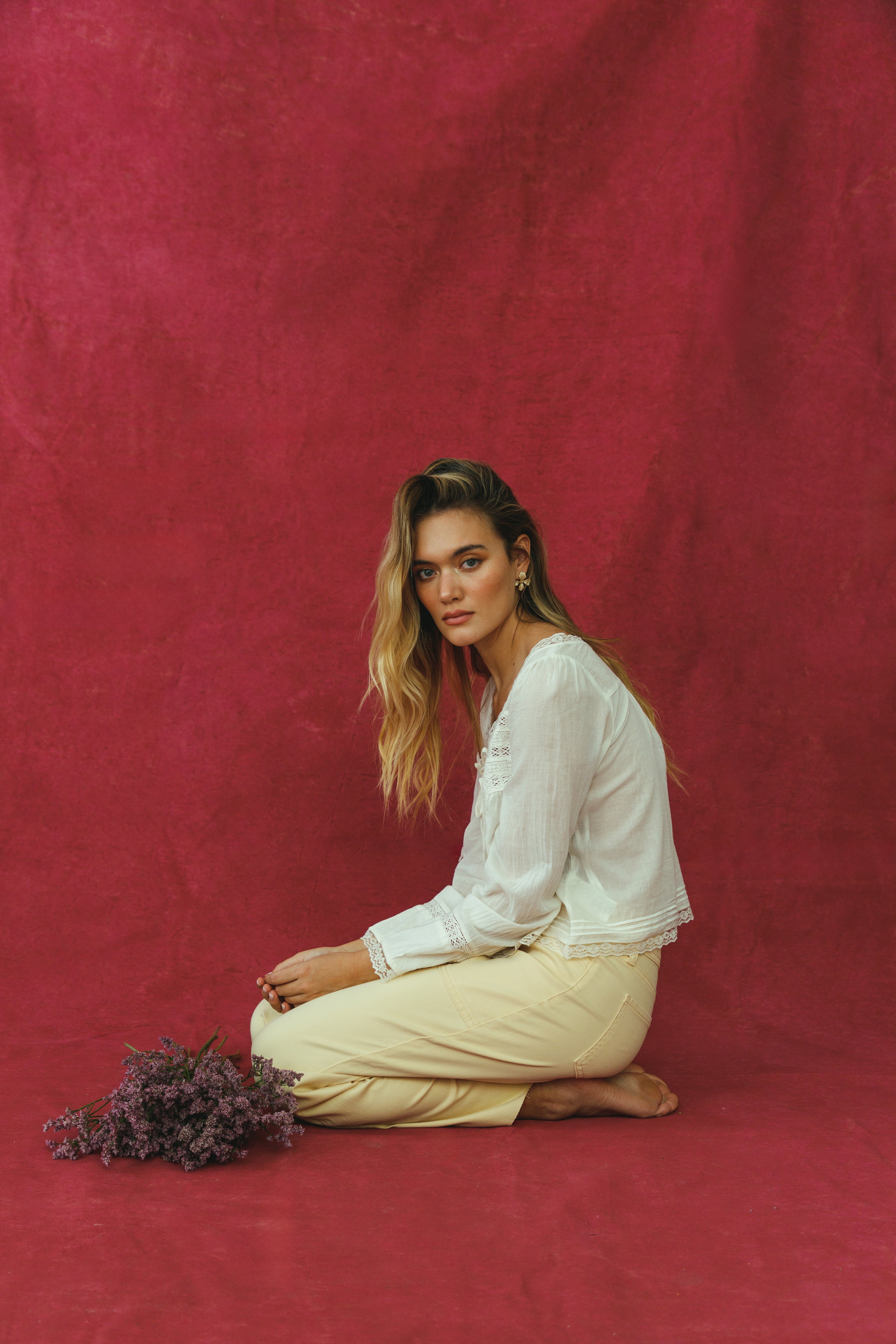 Woman in a white shirt and beige pants sitting on a red background with a bundle of flowers.