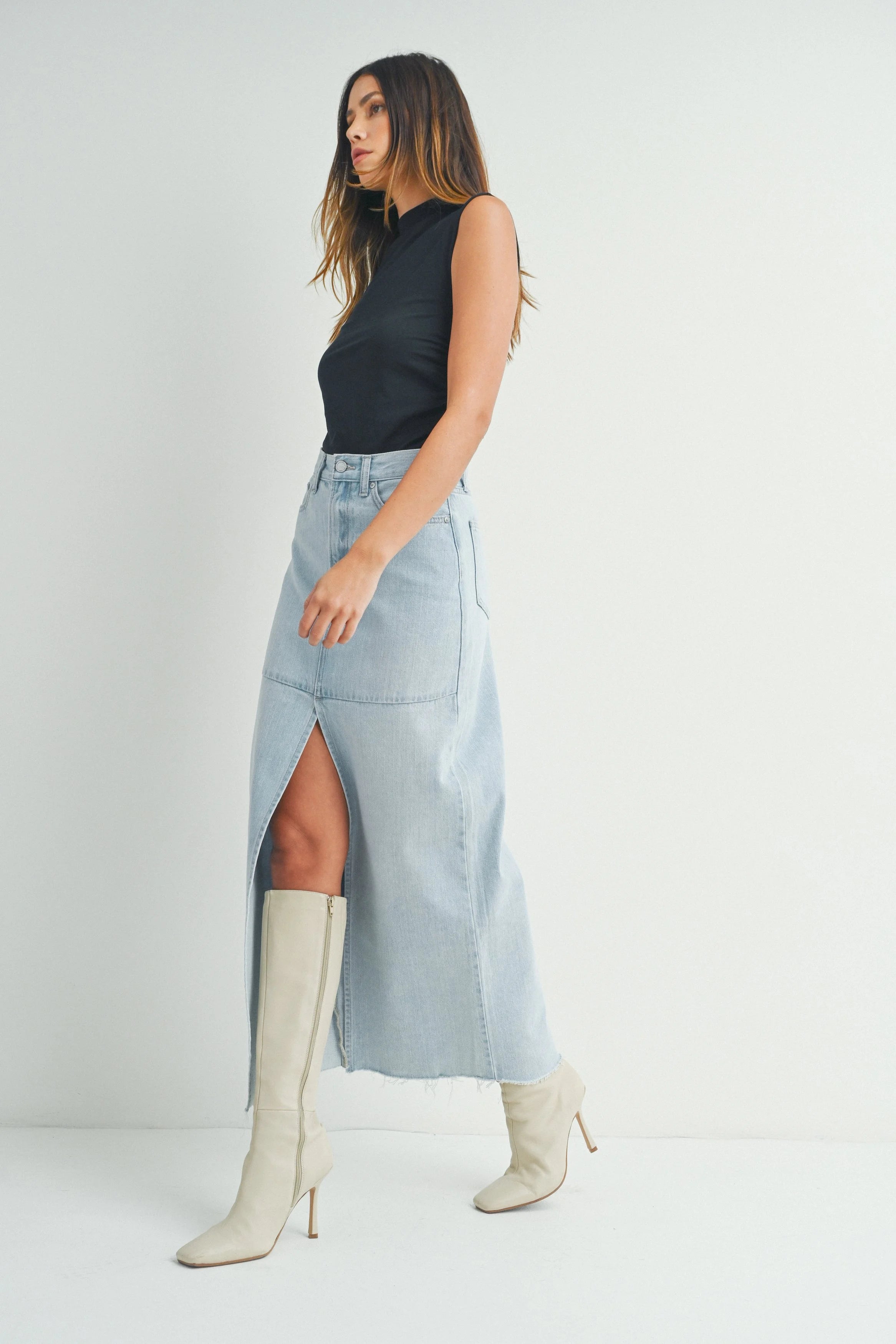 Woman wearing a black sleeveless top, light blue denim skirt, and white knee-high boots on a plain background