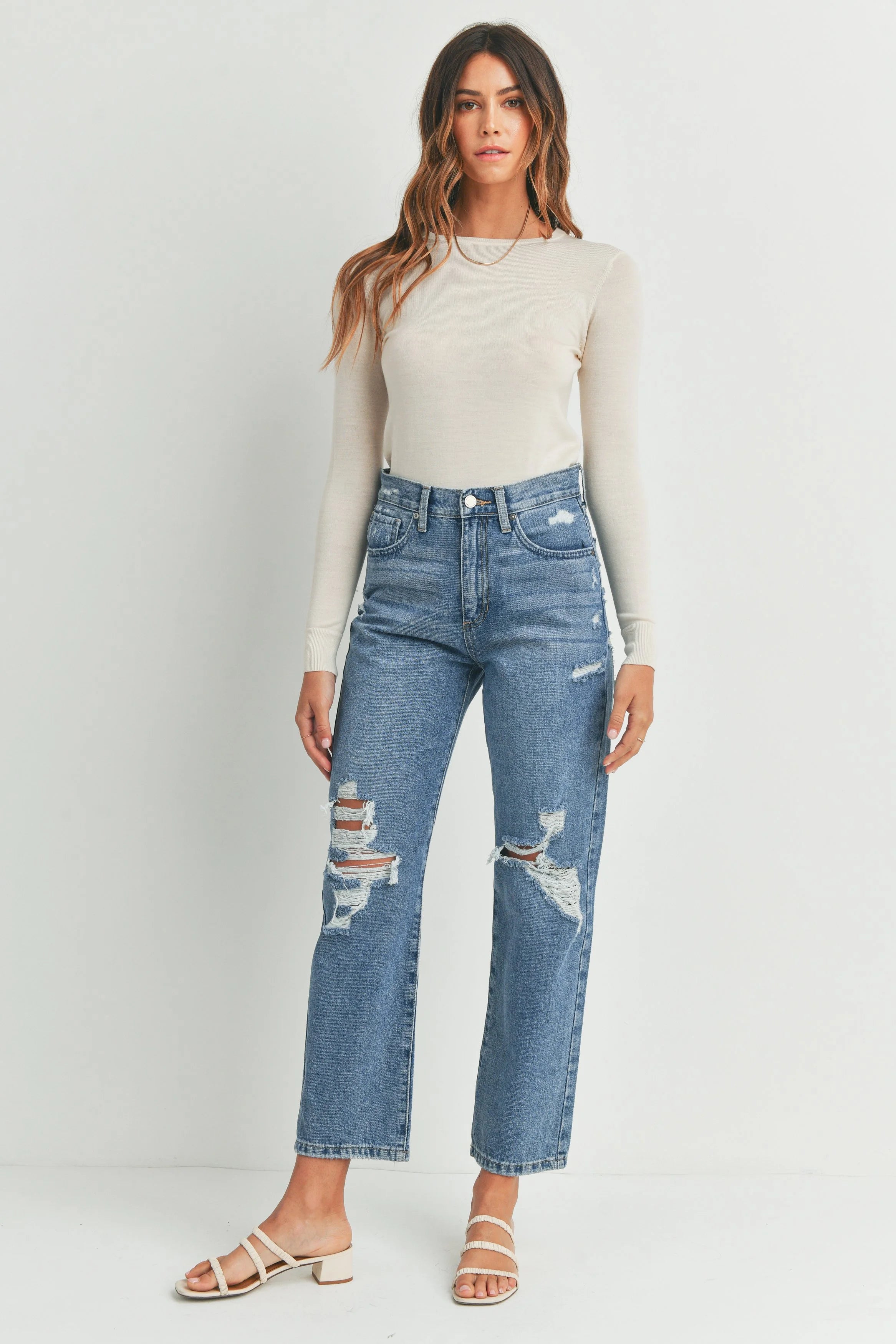 Woman wearing a white long-sleeve top and blue distressed jeans on a plain background