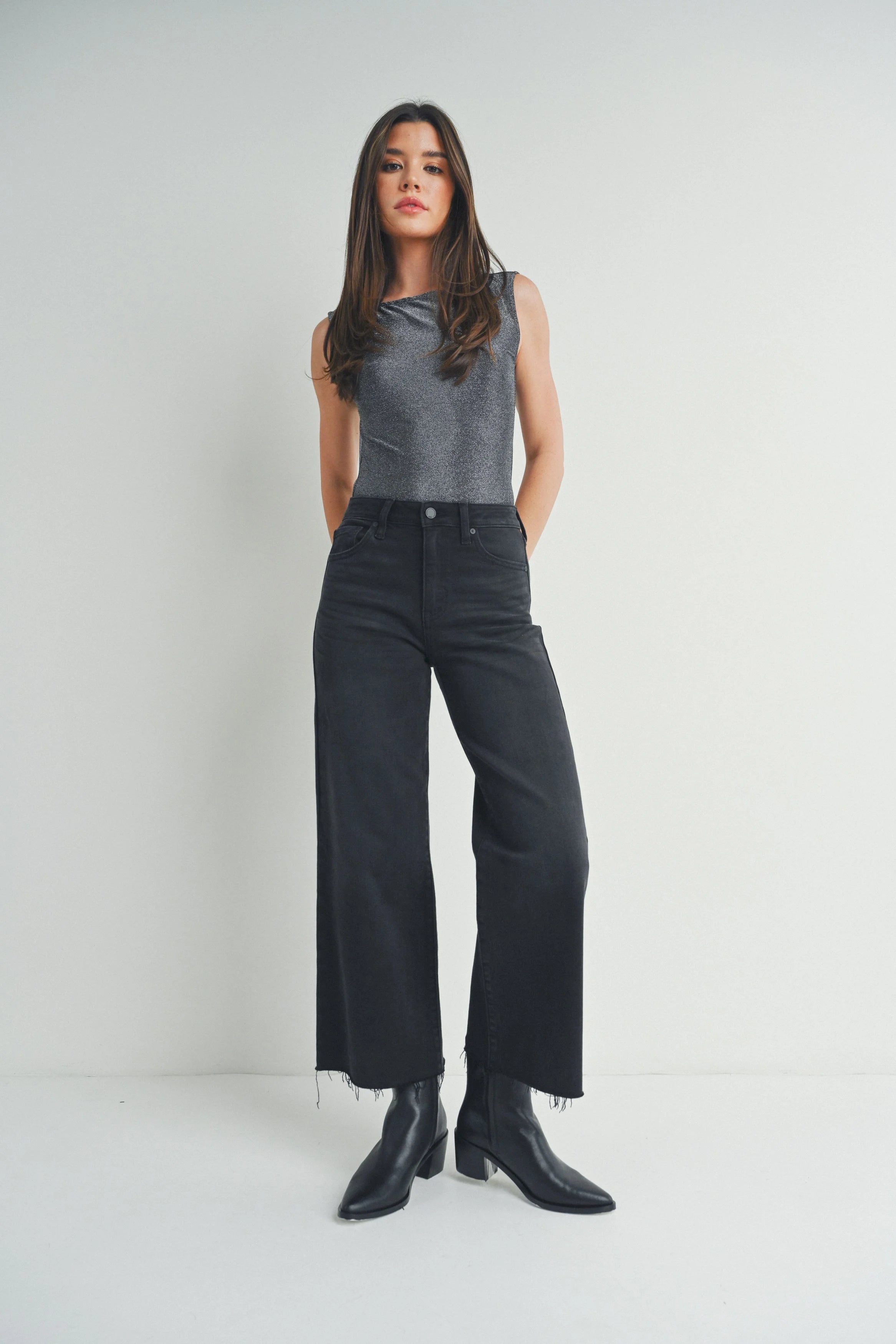 Woman wearing a gray sleeveless top and black jeans with black boots on a white background