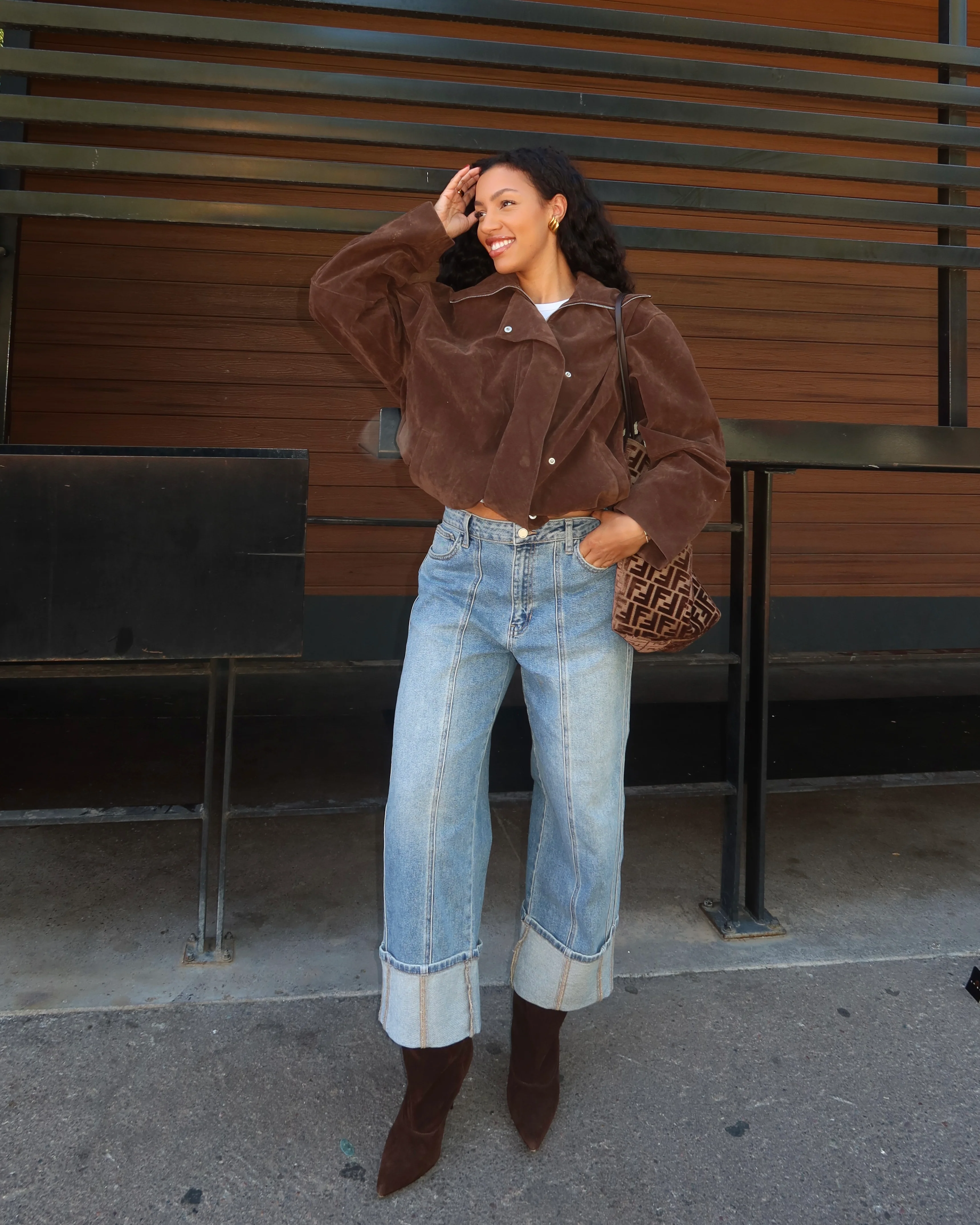 Woman wearing a brown jacket and blue jeans with a handbag, standing against a wooden wall.